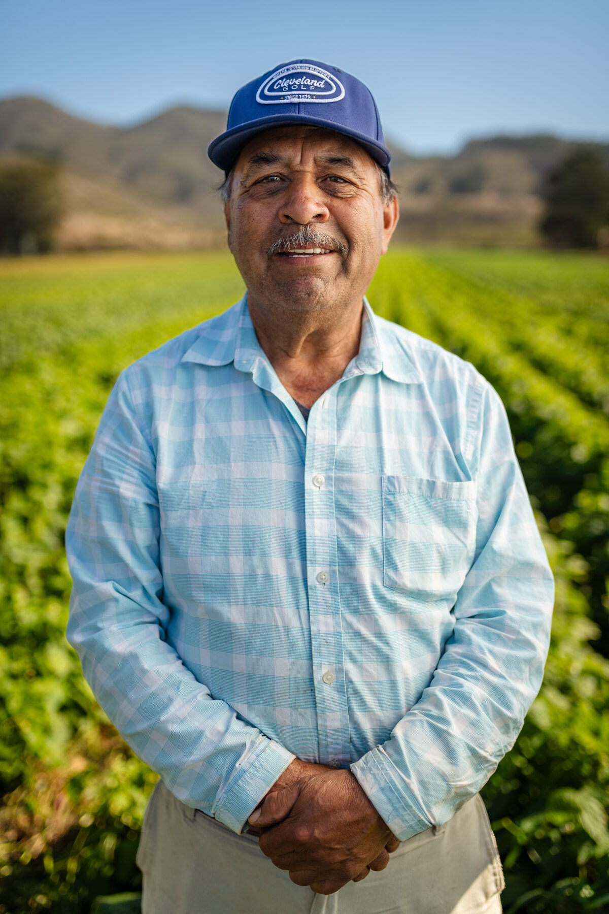 Senior farmworker in a light blue plaid shirt, smiling in the field