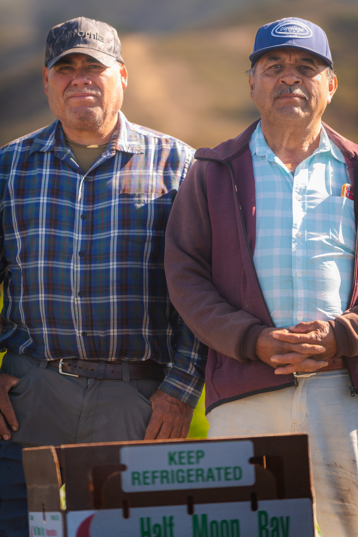 Two senior farmworkers standing together with a box of Half Moon Bay produce