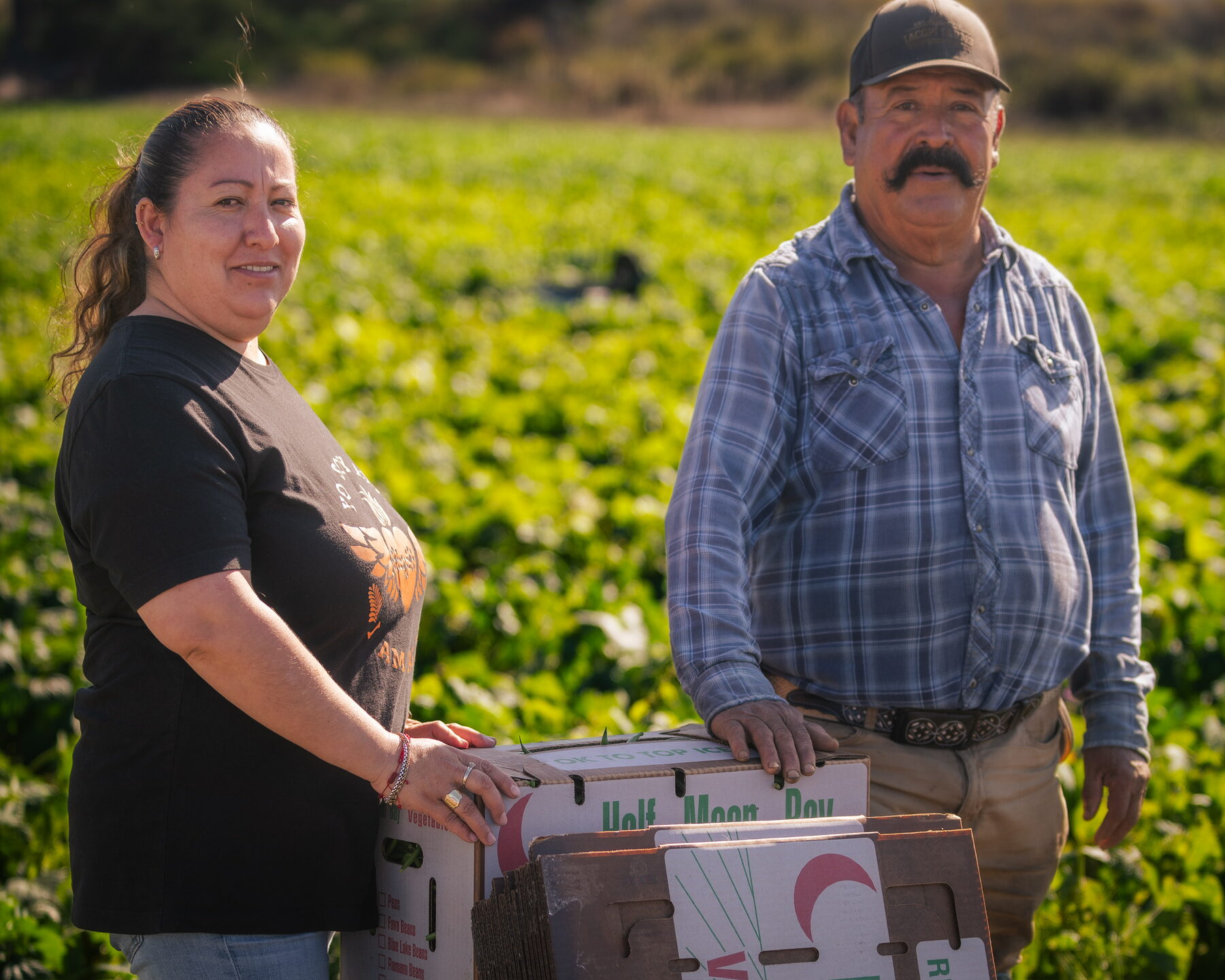 Two senior farmworkers smiling together with a box of Half Moon Bay produce